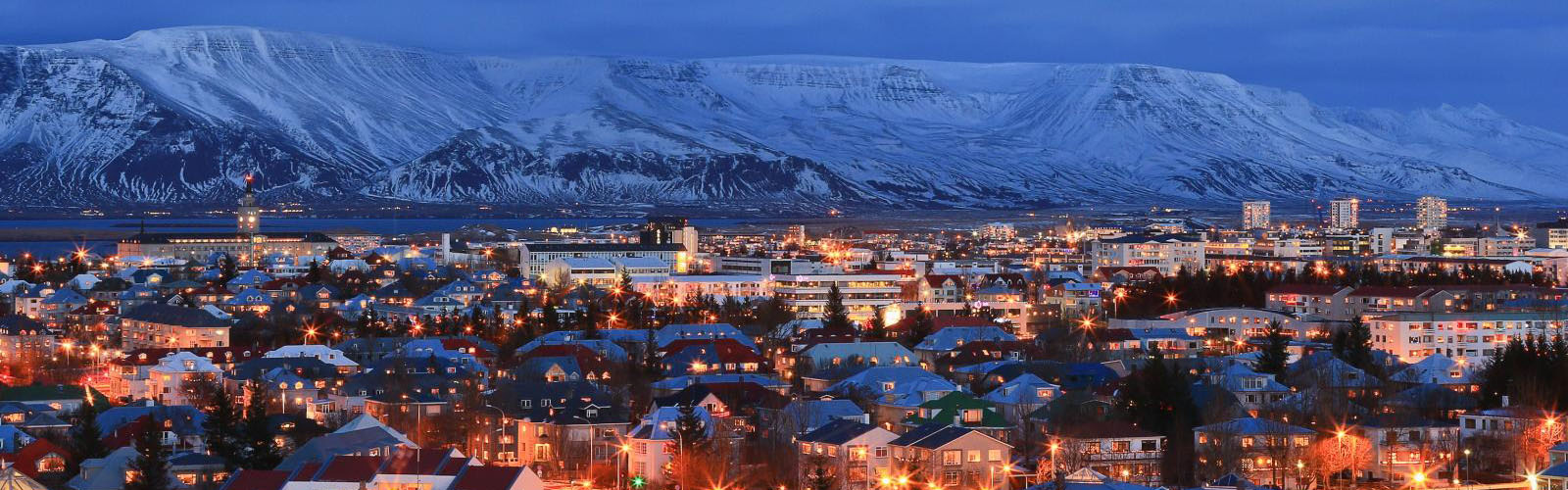 The city of Reykjavik at night, full of buildings with pinpricks of light. A looming, snow-covered mountain is visible behind the buildings. 